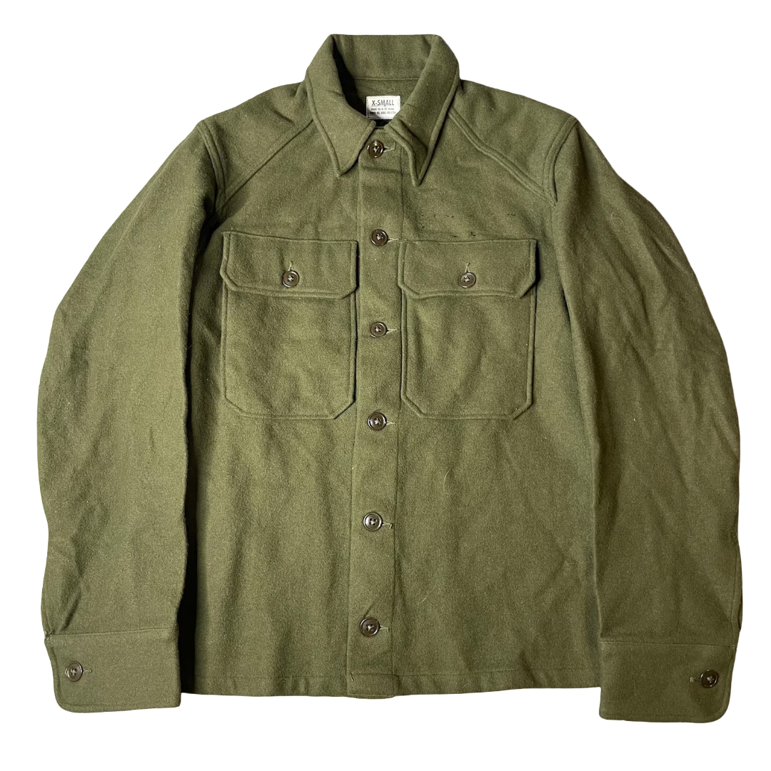 A military-style camouflage jacket laid flat on a white fabric surface. The pattern is a mix of green, brown, black, and tan in a digital camouflage design, with flap chest pockets and a collar.